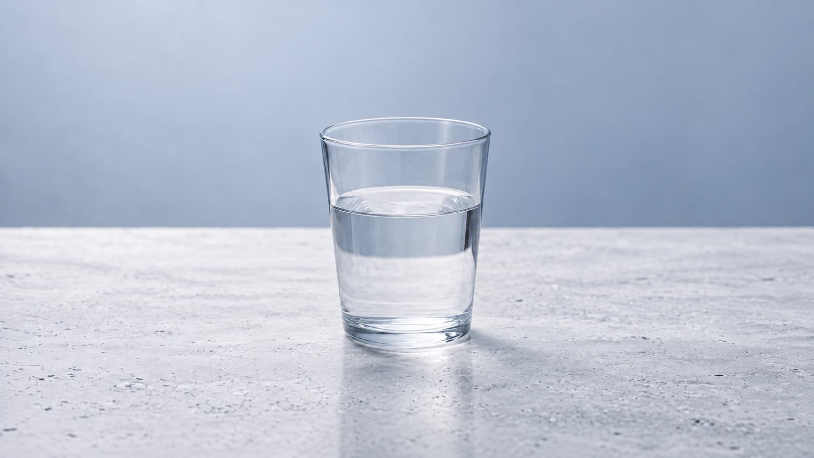 A single glass half-filled with clear water on pale stone against a blue-gray background