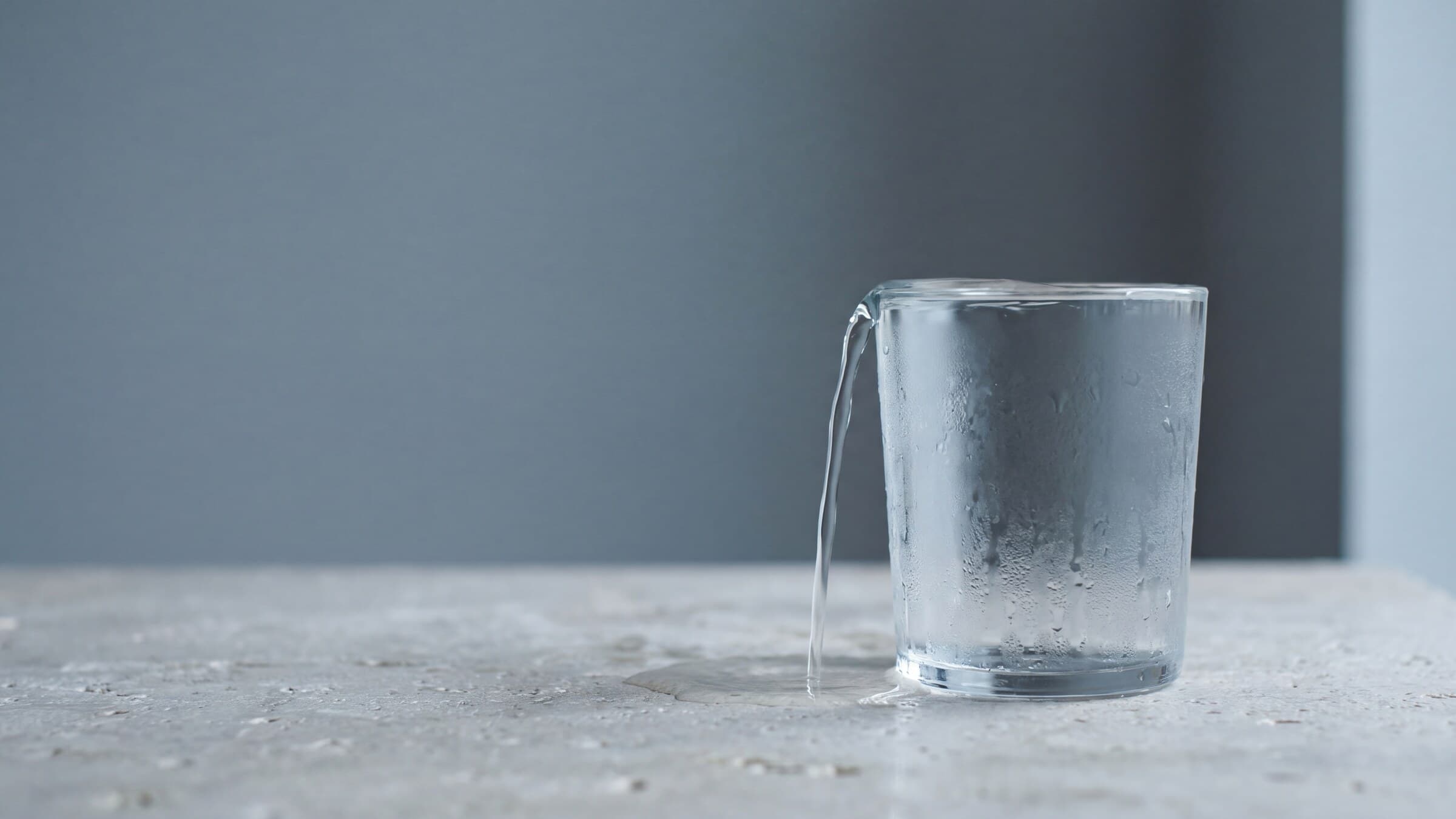A clear drinking glass overflowing onto pale stone against a blue-gray background