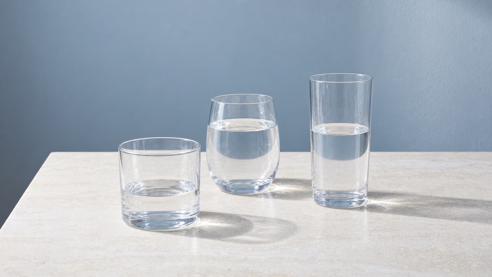 Three differently shaped drinking glasses with varying water levels on pale stone