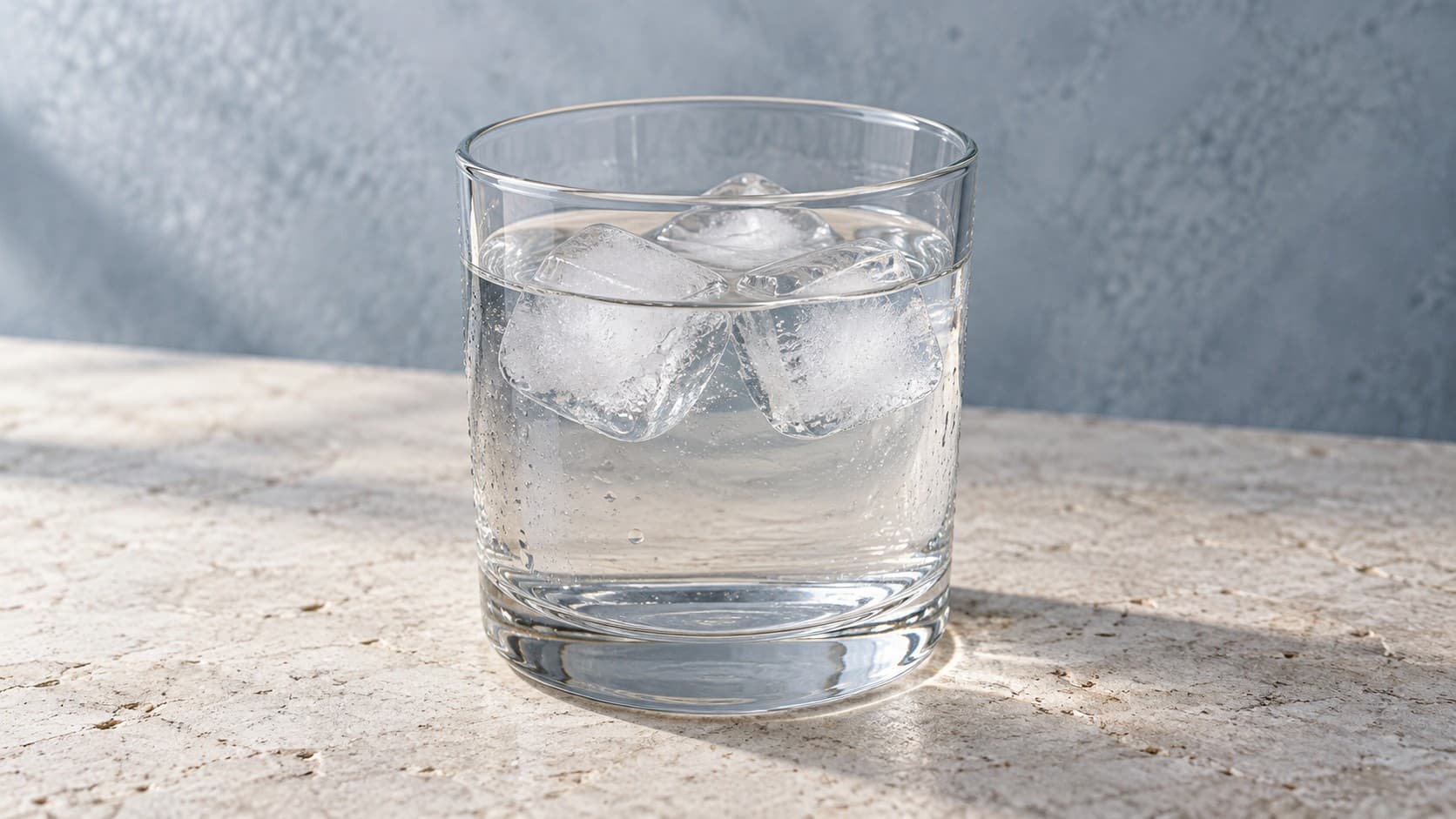 Three ice cubes floating in a clear glass of water on pale limestone against a blue-gray wall