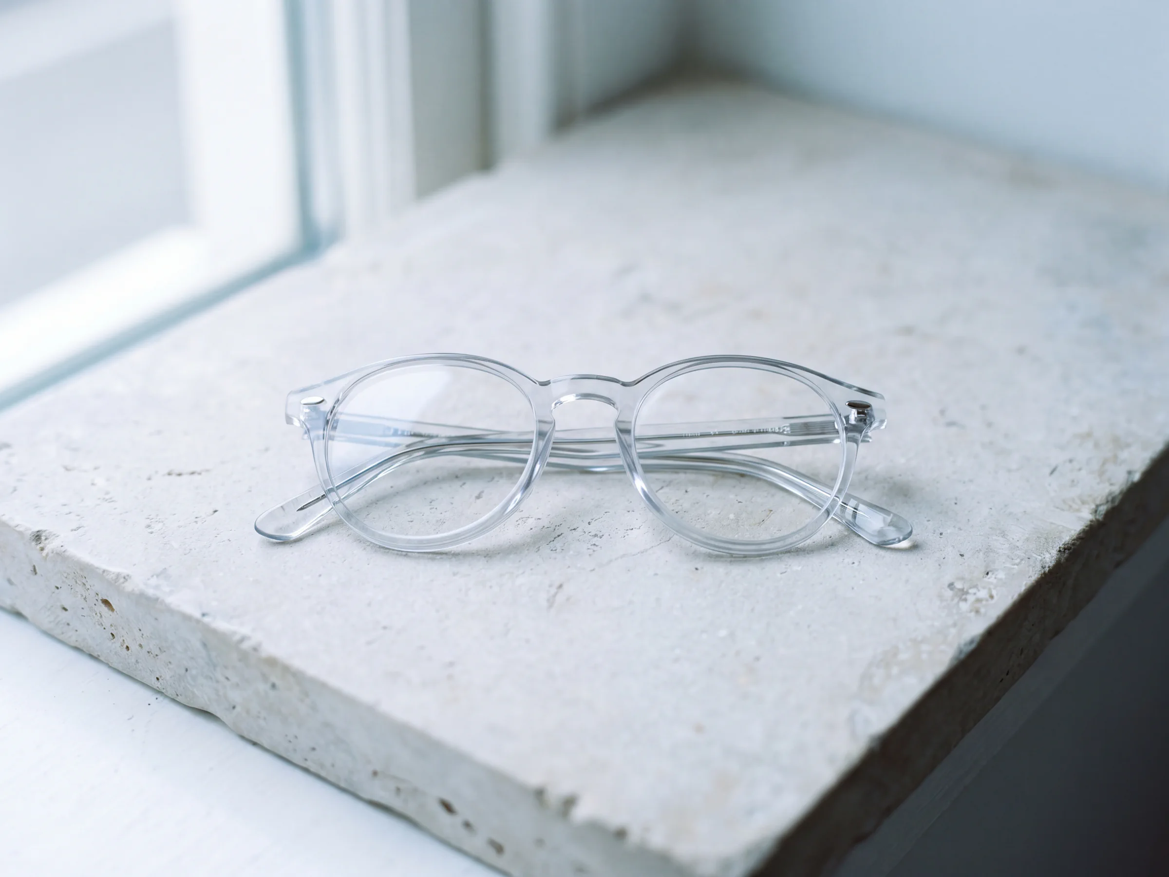 A pair of clear eyeglasses resting on a pale stone surface in soft window light