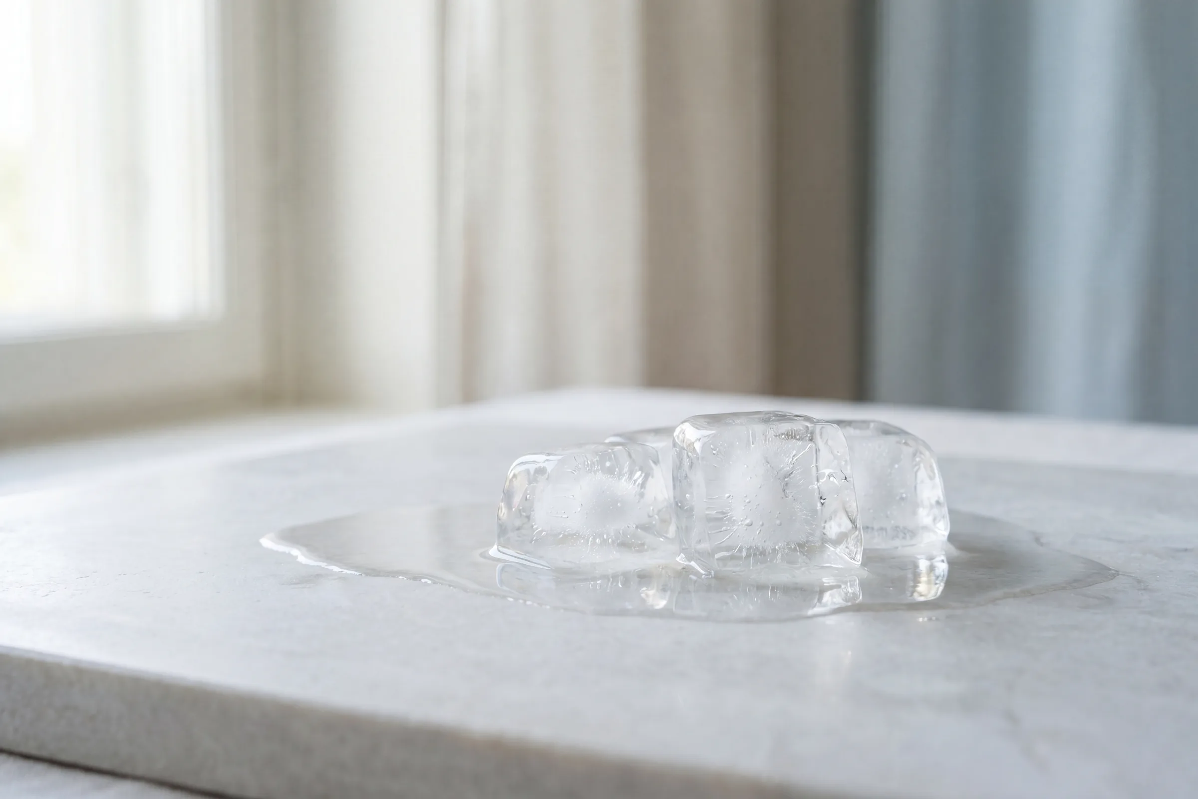 A melting ice cube in a small pool of water on a pale marble surface in soft window light